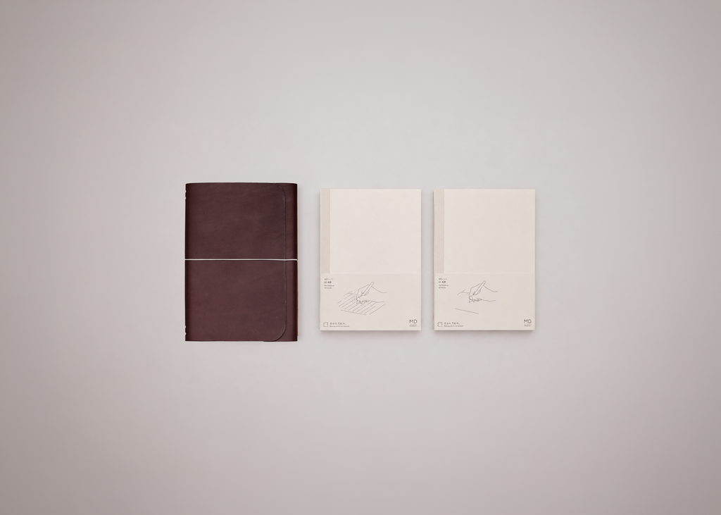One leather journal cover in dark brown next to two white notebook inserts against a light grey background