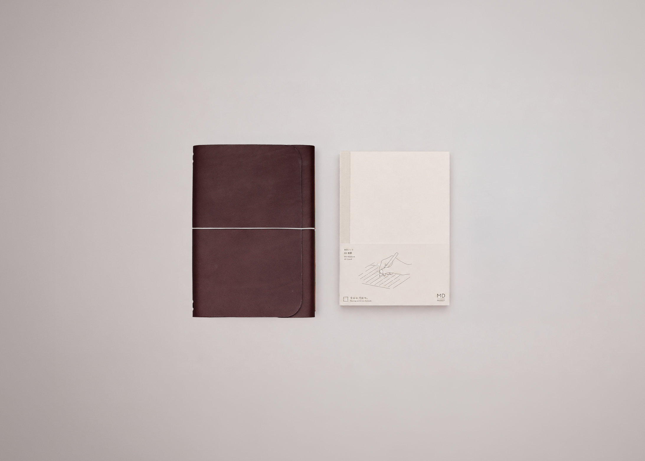 A dark brown leather-bound notebook with a thin elastic band closure placed next to a small white paper notebook insert set against a clean light grey background.