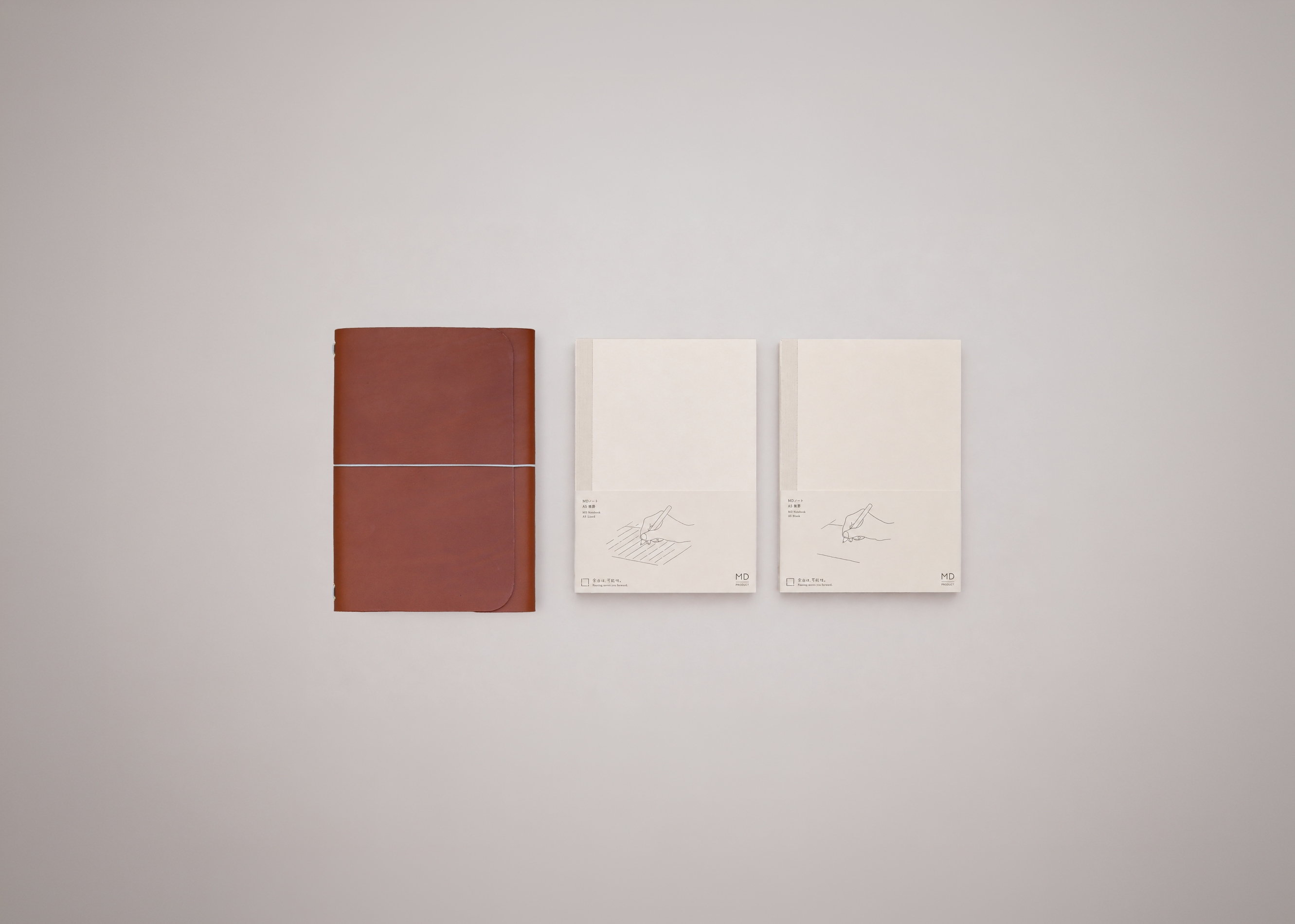 Cognac leather journal with a white elastic closure horizontally across the centre, next to two white notebooks on a light grey background