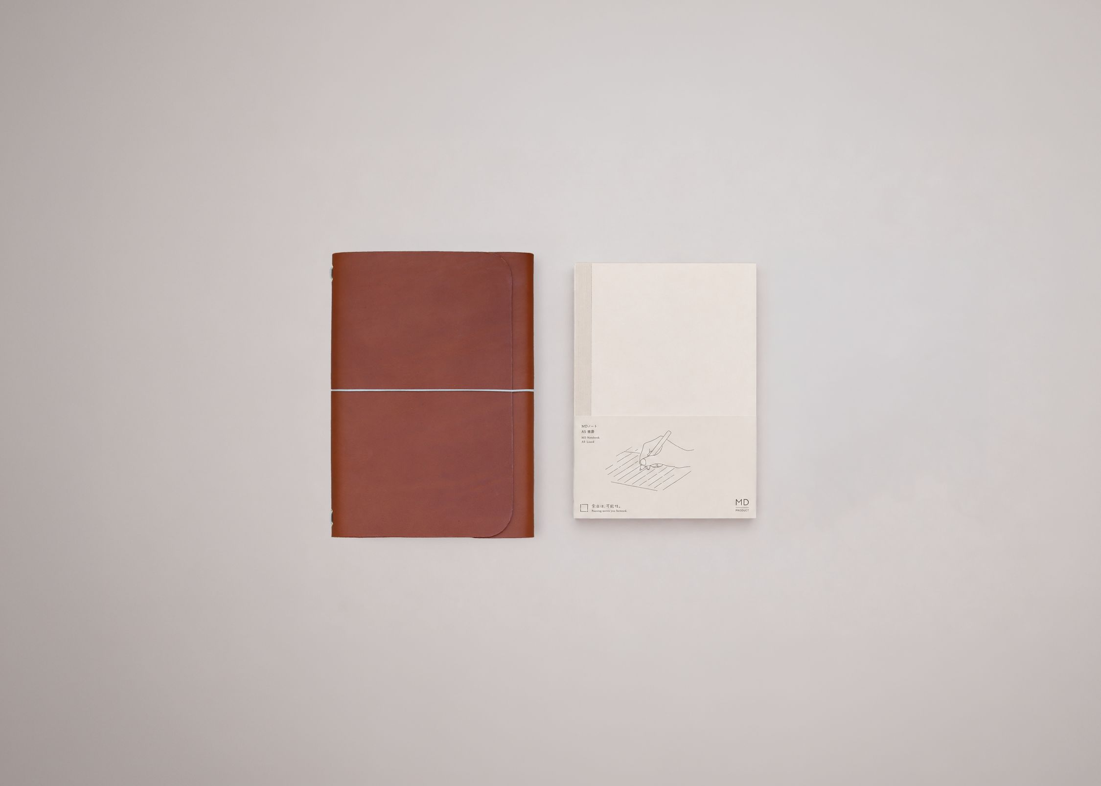 A brown leather-bound notebook with a horizontal centre elastic sits next to a white paper notebook with a minimalist line drawing on a light gray background.