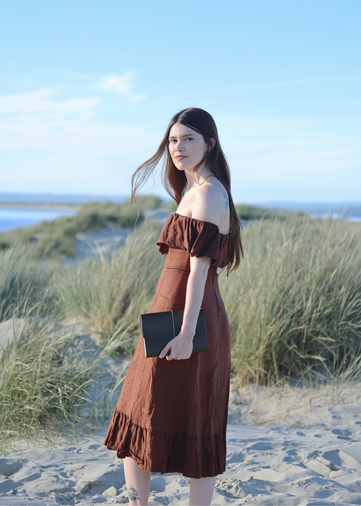 A woman stands on a sandy beach dune wearing a brown off-the-shoulder midi dress and holding an olive green leather journal, with sea grass and the ocean in the background under a blue sky.