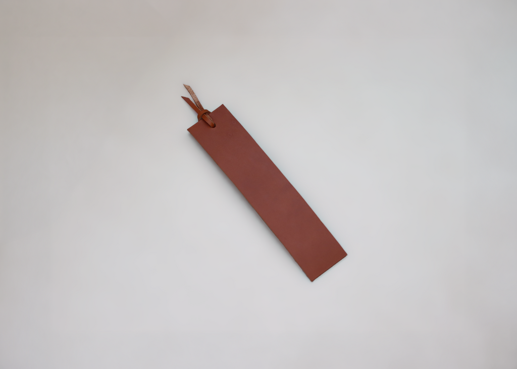 A minimalist brown leather bookmark with a small leather loop and tassel, photographed on a clean, light grey background.