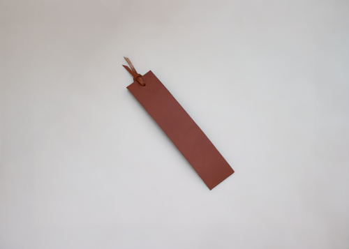A minimalist brown leather bookmark with a small leather loop and tassel, photographed on a clean, light grey background.