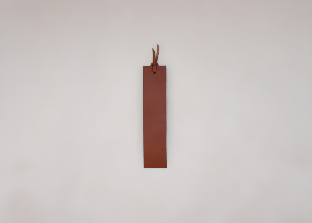A minimalist brown leather bookmark with a small leather loop at the top, shown against a plain, light grey background.