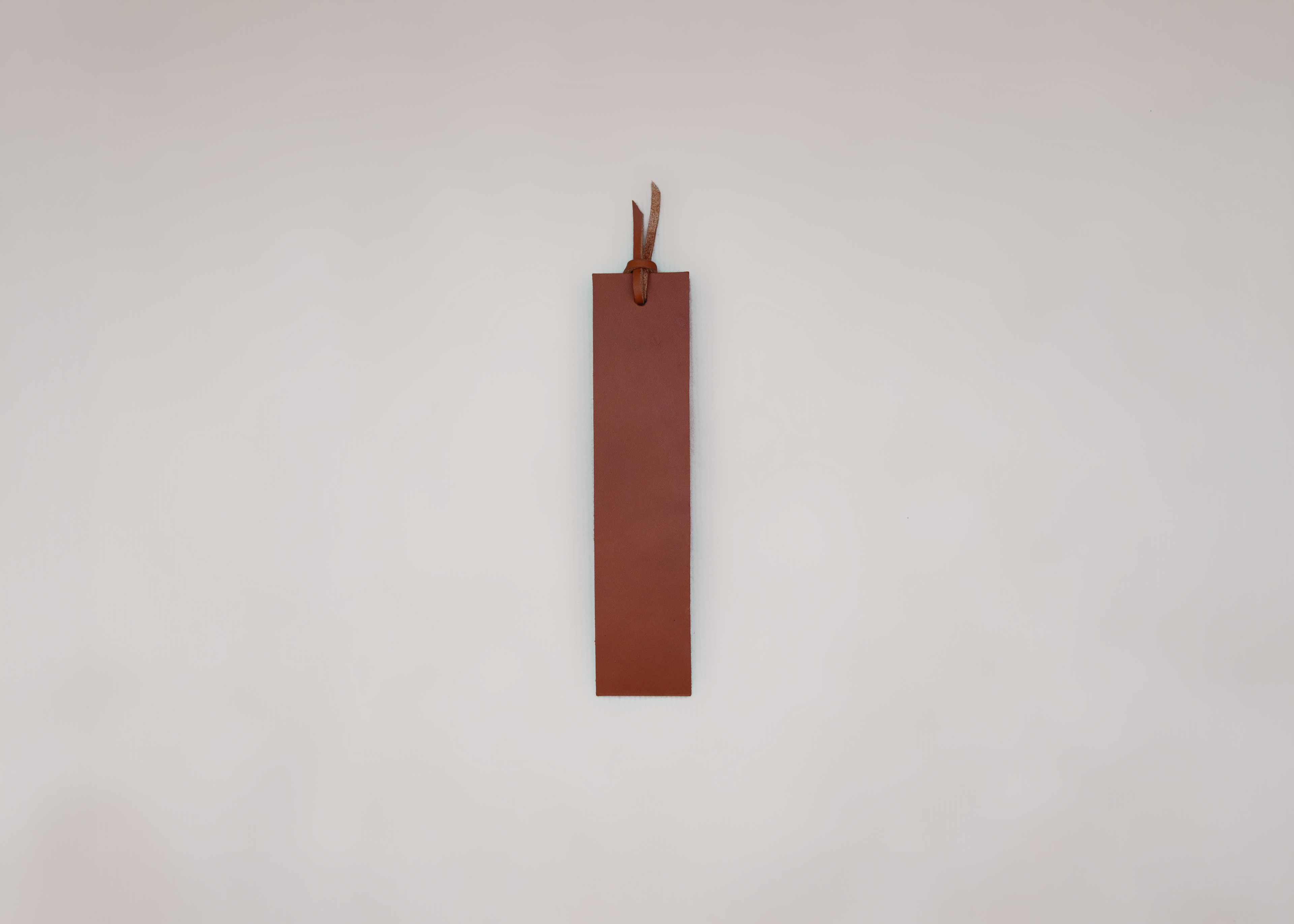A minimalist brown leather bookmark with a small leather loop at the top, shown against a plain, light grey background.