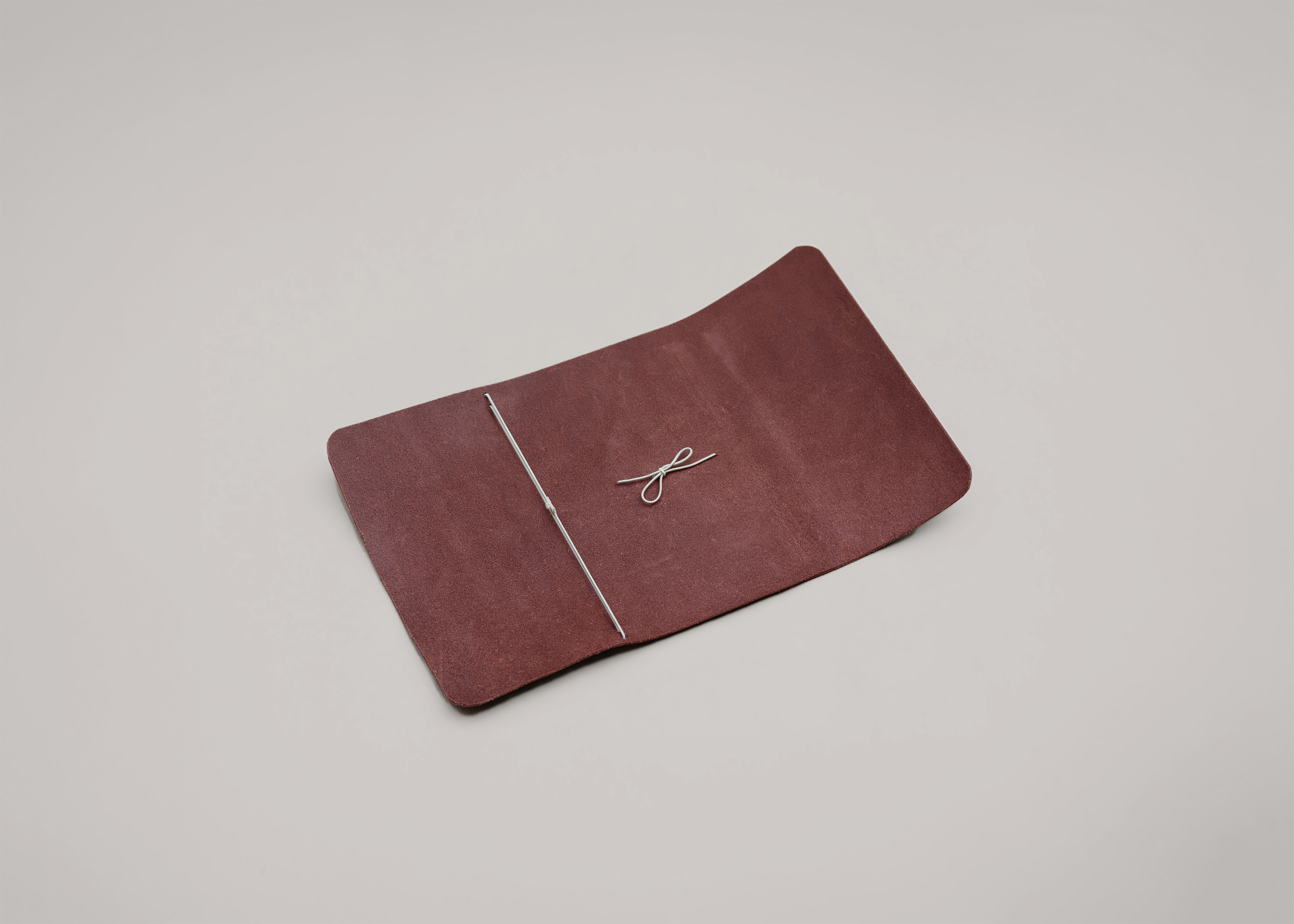 A burgundy leather notebook holder with a white elastic accent, photographed on a neutral light grey background.