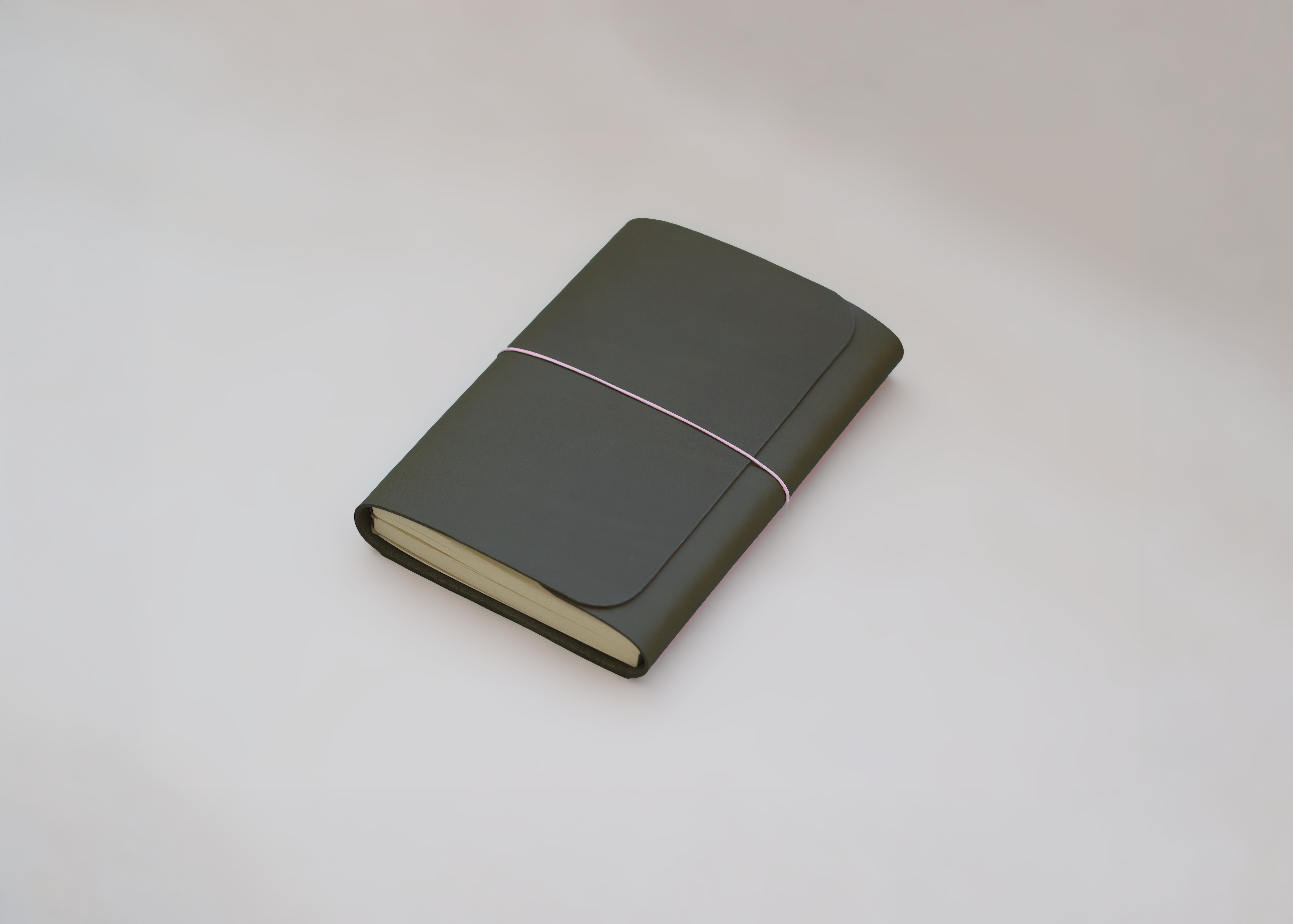 A dark olive green leather-bound notebook with a thin pink elastic band closure and cream-colored pages, photographed on a neutral light grey background.