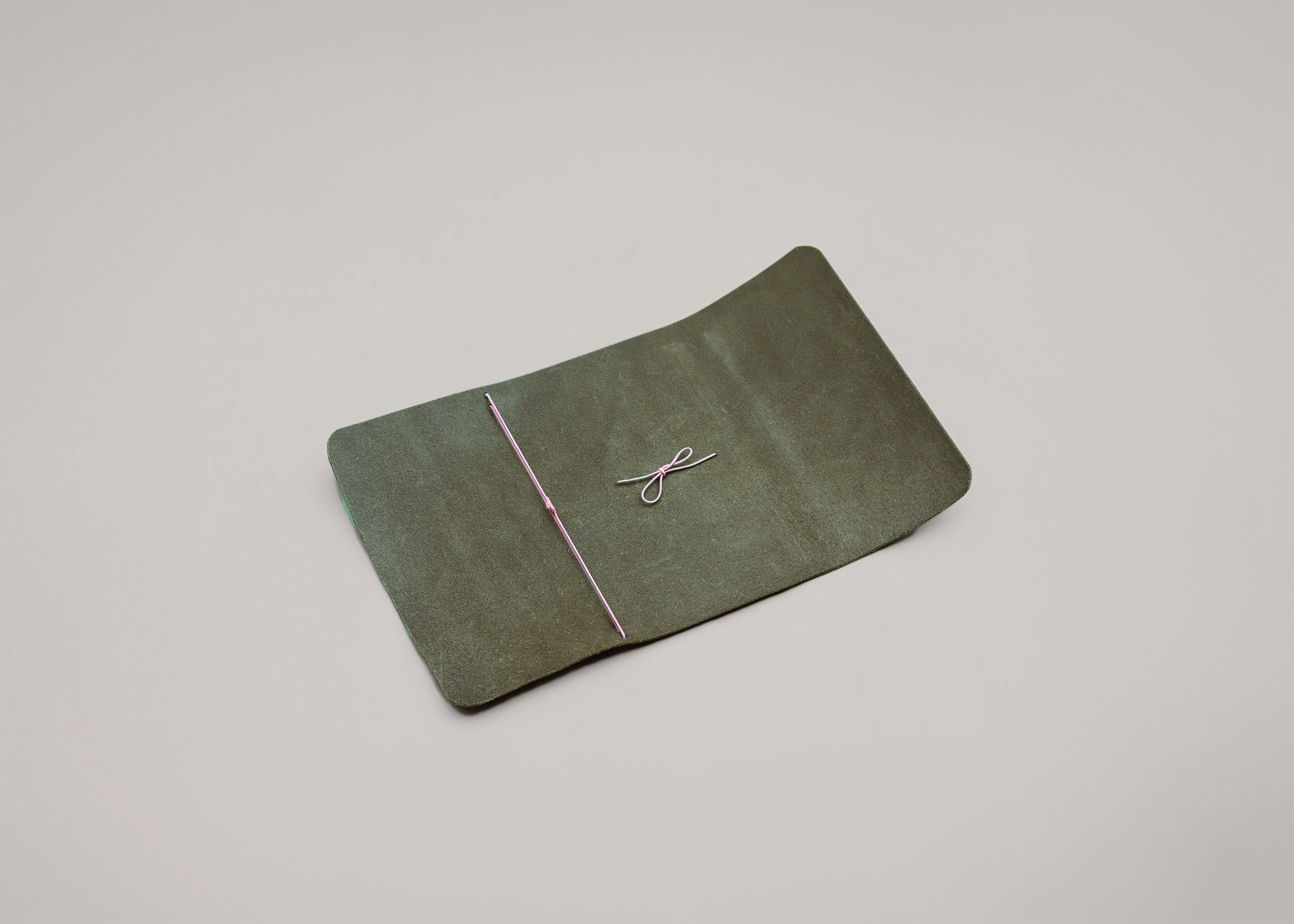 An olive green leather journal holder with a thin pink string tied in a bow across the front, photographed on a neutral light grey background.