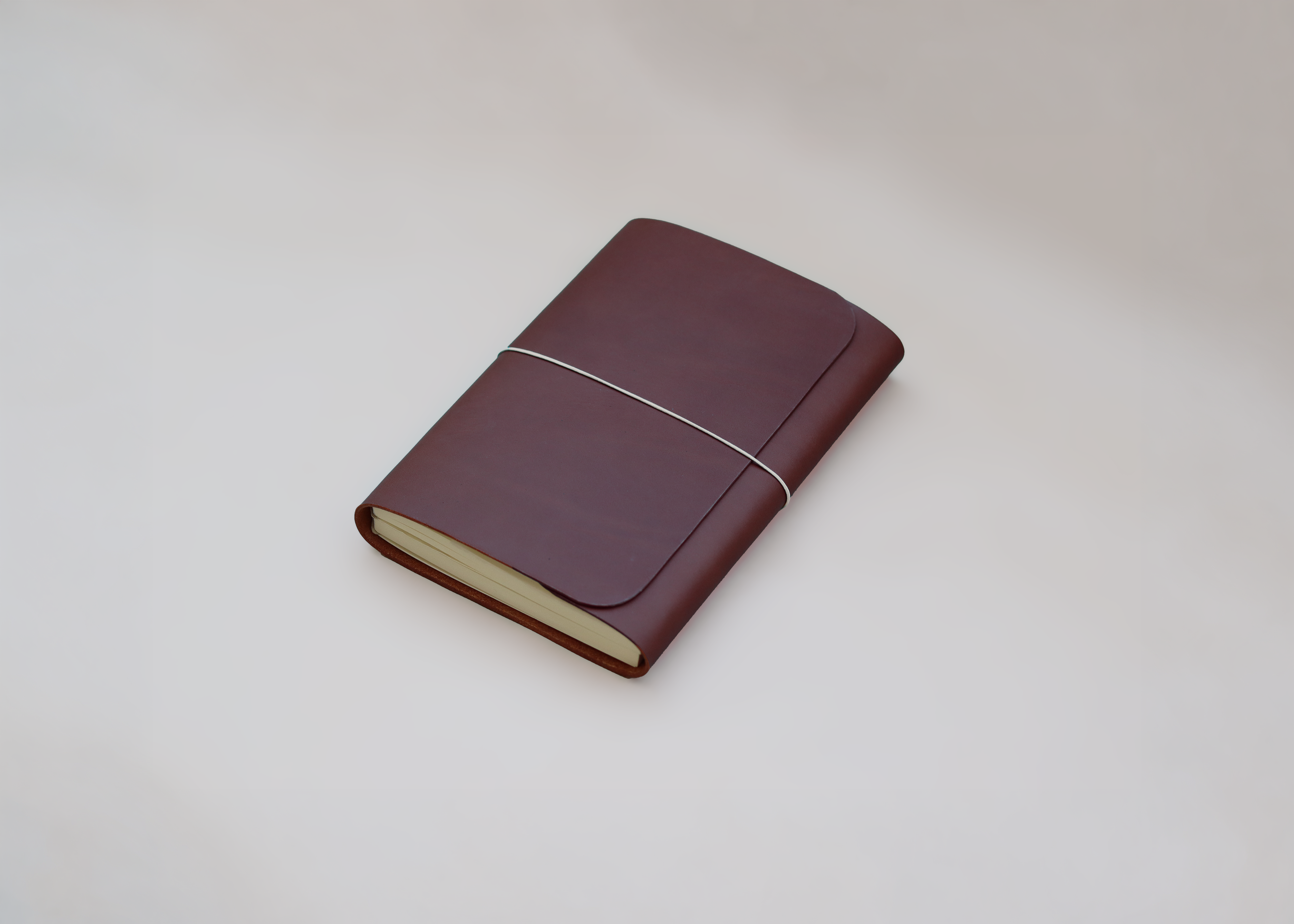 A dark brown leather journal with a white elastic band closure and cream-colored pages, shown on a neutral light grey background.