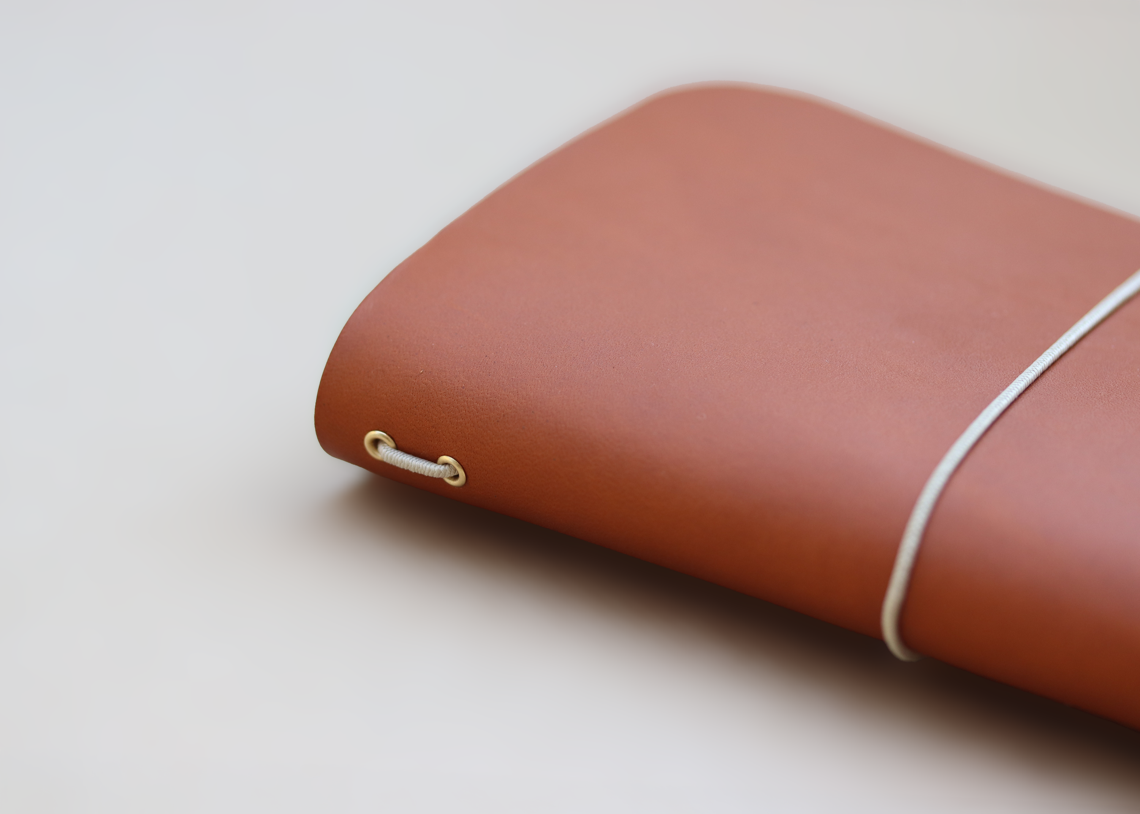 A close-up of a terracotta brown leather notebook cover with a white elastic band and brass grommet details, resting on a light beige surface.