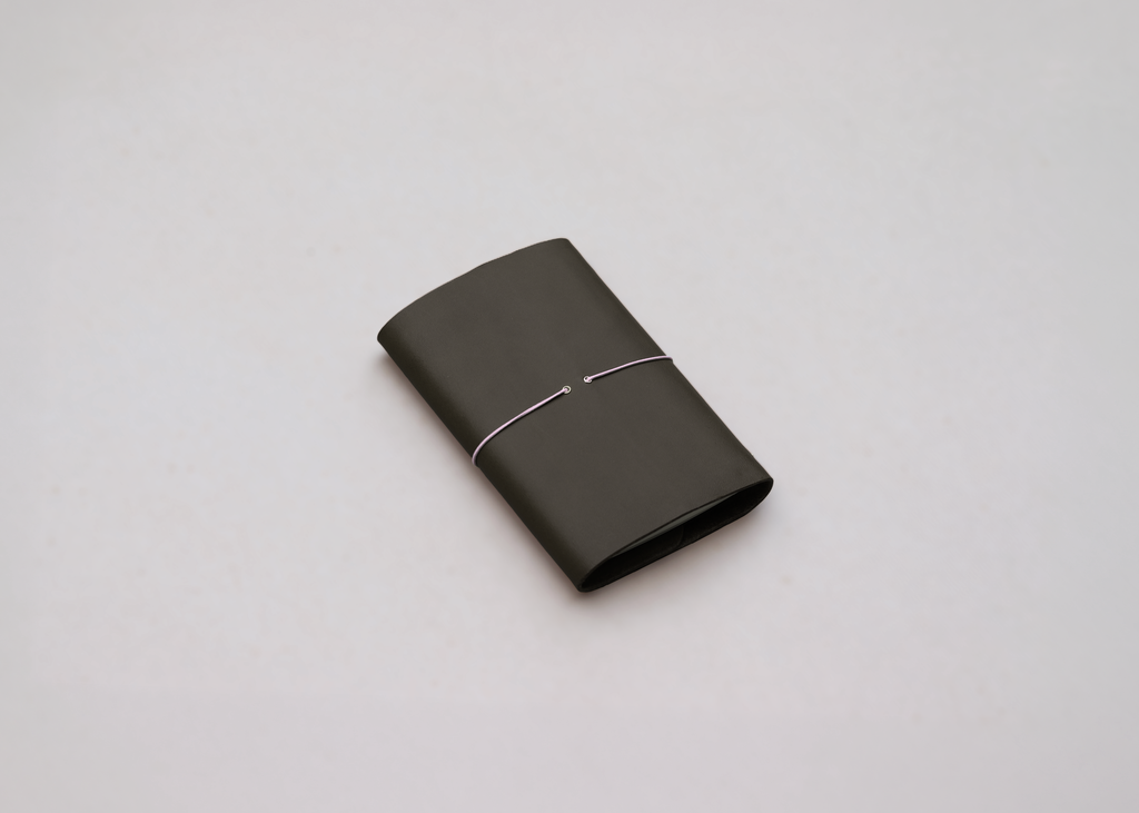 An olive green leather journal with a slim pink elastic around the centre, with brass detailing photographed against a light grey background