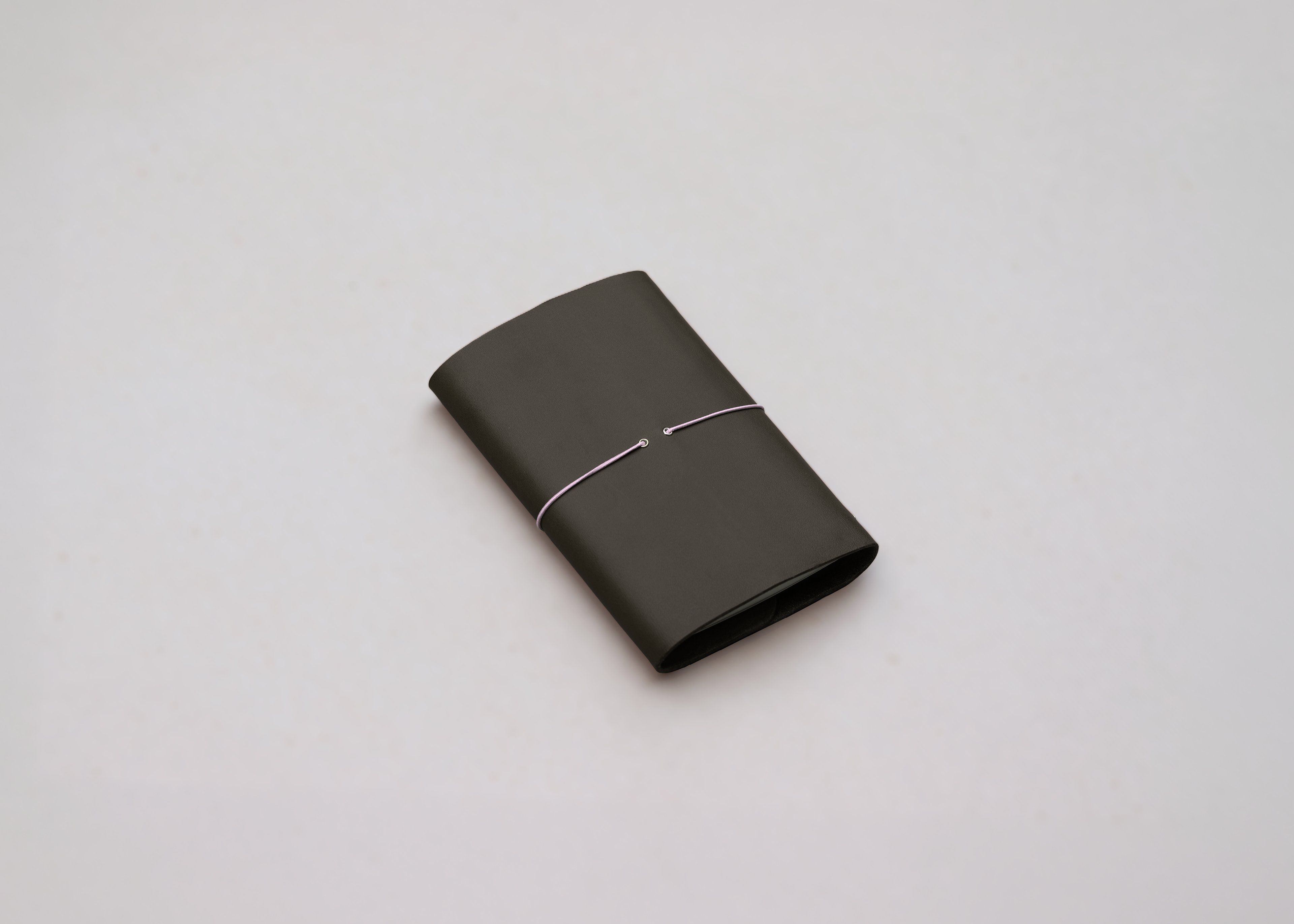 An olive green leather journal with a slim pink elastic around the centre, with brass detailing photographed against a light grey background