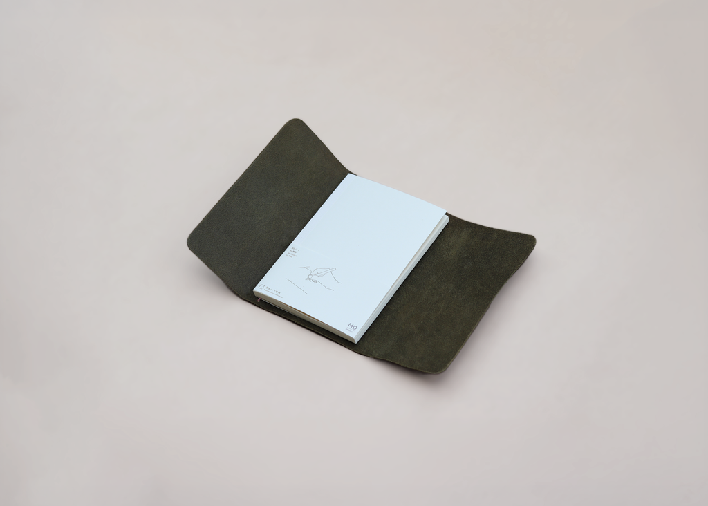 A dark olive green leather notebook cover holding a white notebook, photographed on a neutral light beige background.
