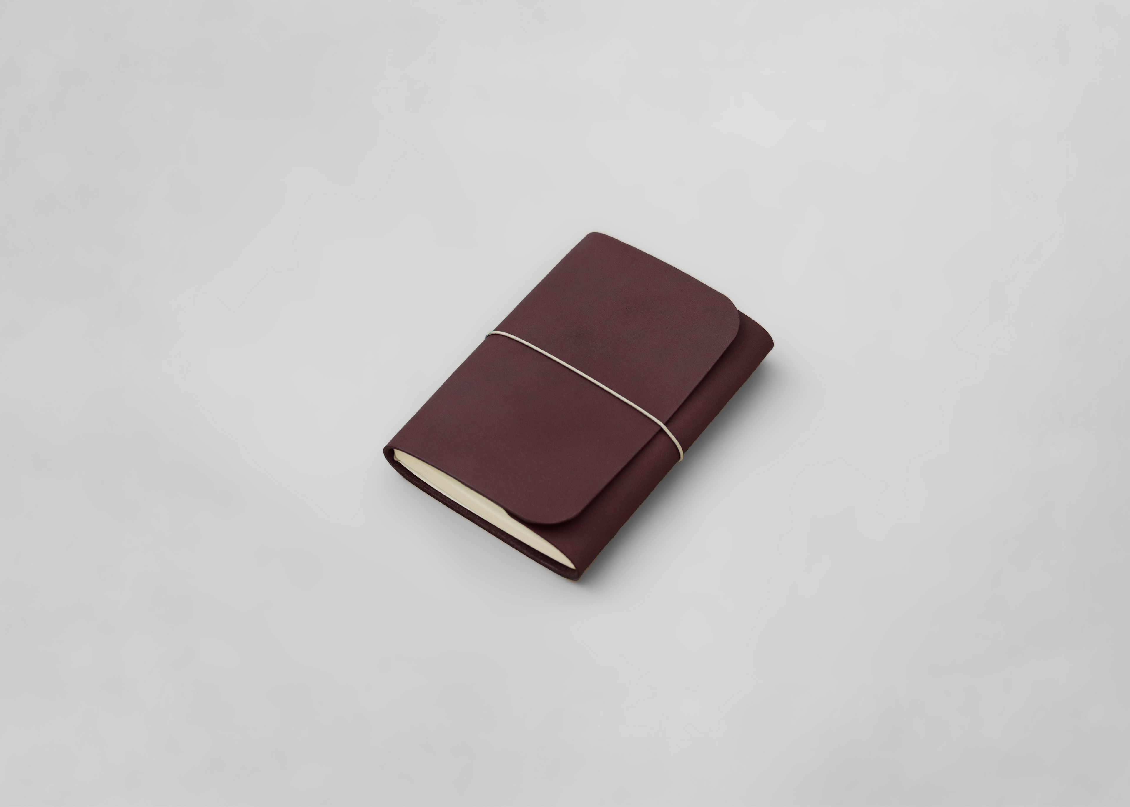 A dark reddish-brown leather journal holder with a flap cover and a thin white elastic band, photographed on a light grey background.