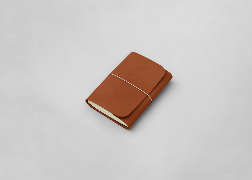 A brown leather journal with a flap cover and a thin white elastic band, photographed on a light grey background.