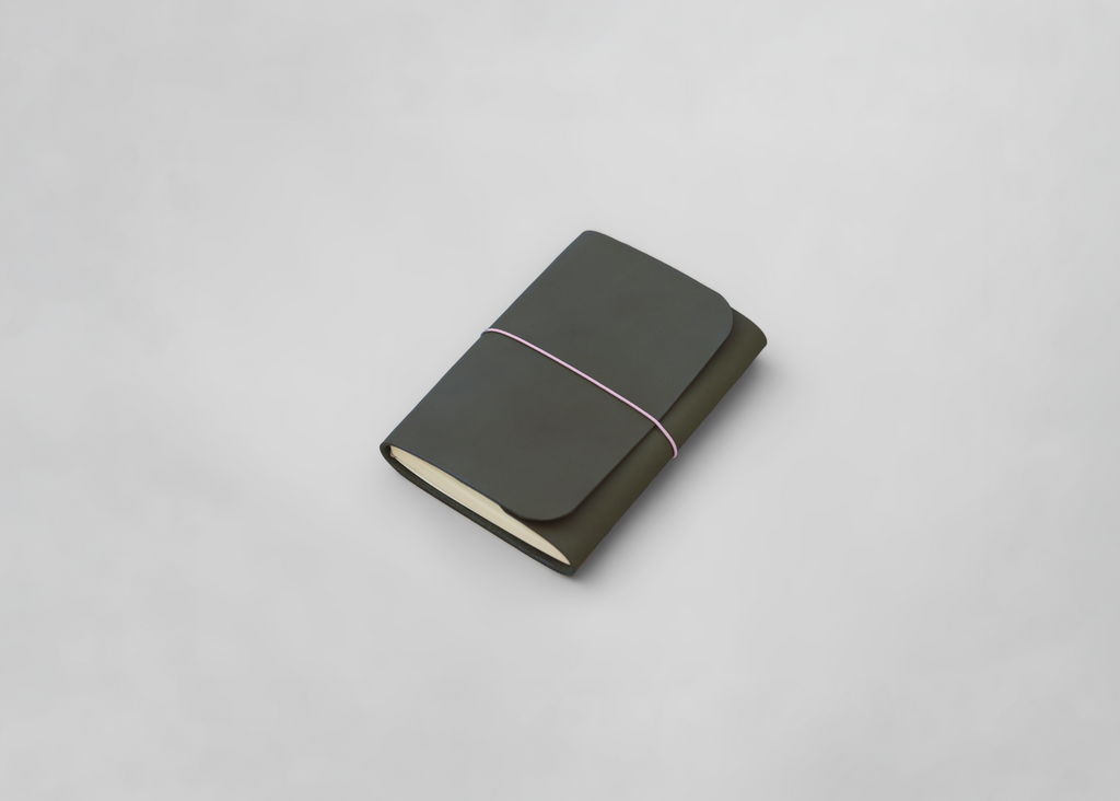 A leather journal in dark olive green with a light pink elastic band and cream-colored pages, photographed on a clean light grey background.
