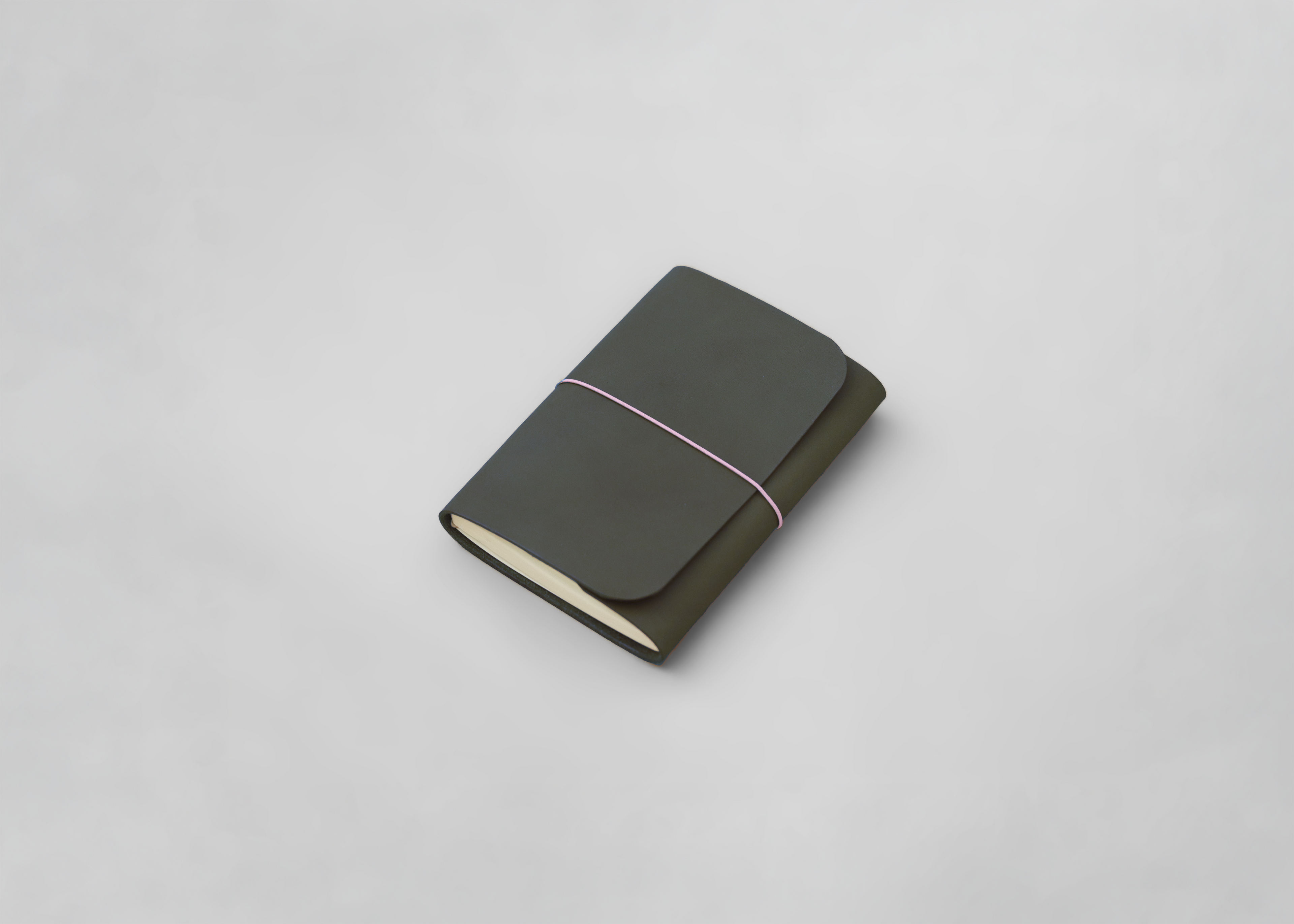 A leather journal in dark olive green with a light pink elastic band and cream-colored pages, photographed on a clean light grey background.