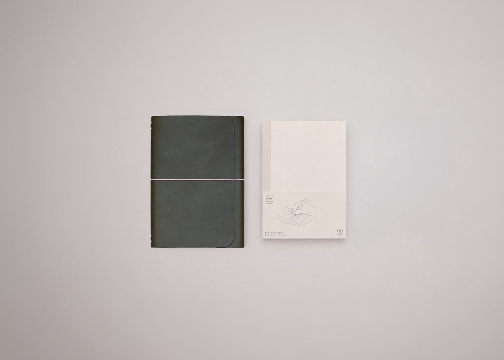A minimalist olive green leather journal displayed next to a white notebook against a light grey background. 